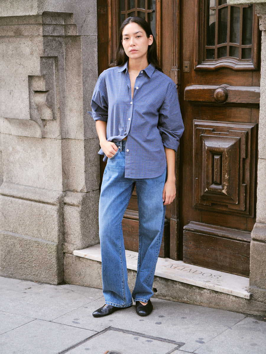 A woman stands on a city sidewalk in front of a stone building and dark wooden door, wearing a blue button-up shirt with rolled sleeves and blue wide-leg jeans.