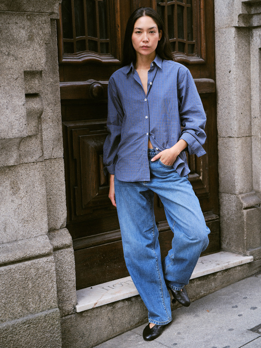 A woman stands on stone steps in front of a dark wooden door, wearing a blue patterned button-up shirt and wide-leg blue jeans.