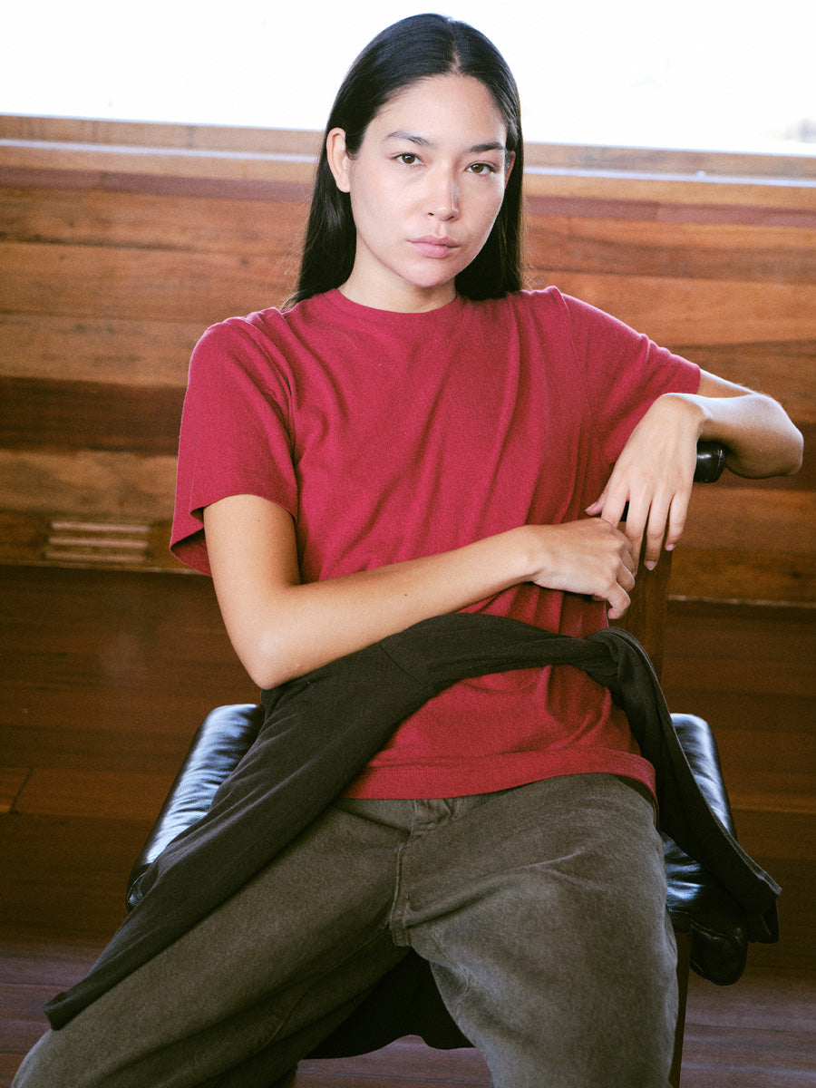 A woman sitting in a red tshirt against a wood wall, with brown pants.