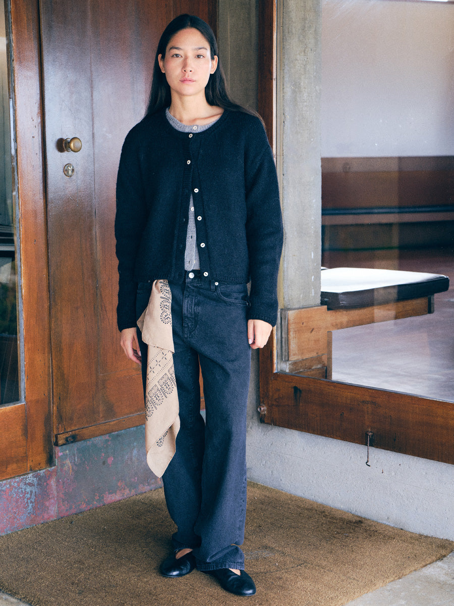A woman stands in a doorway wearing a black button-front cardigan over a gray top, dark wide-leg jeans, and black flats, with a beige patterned scarf draped over one leg.