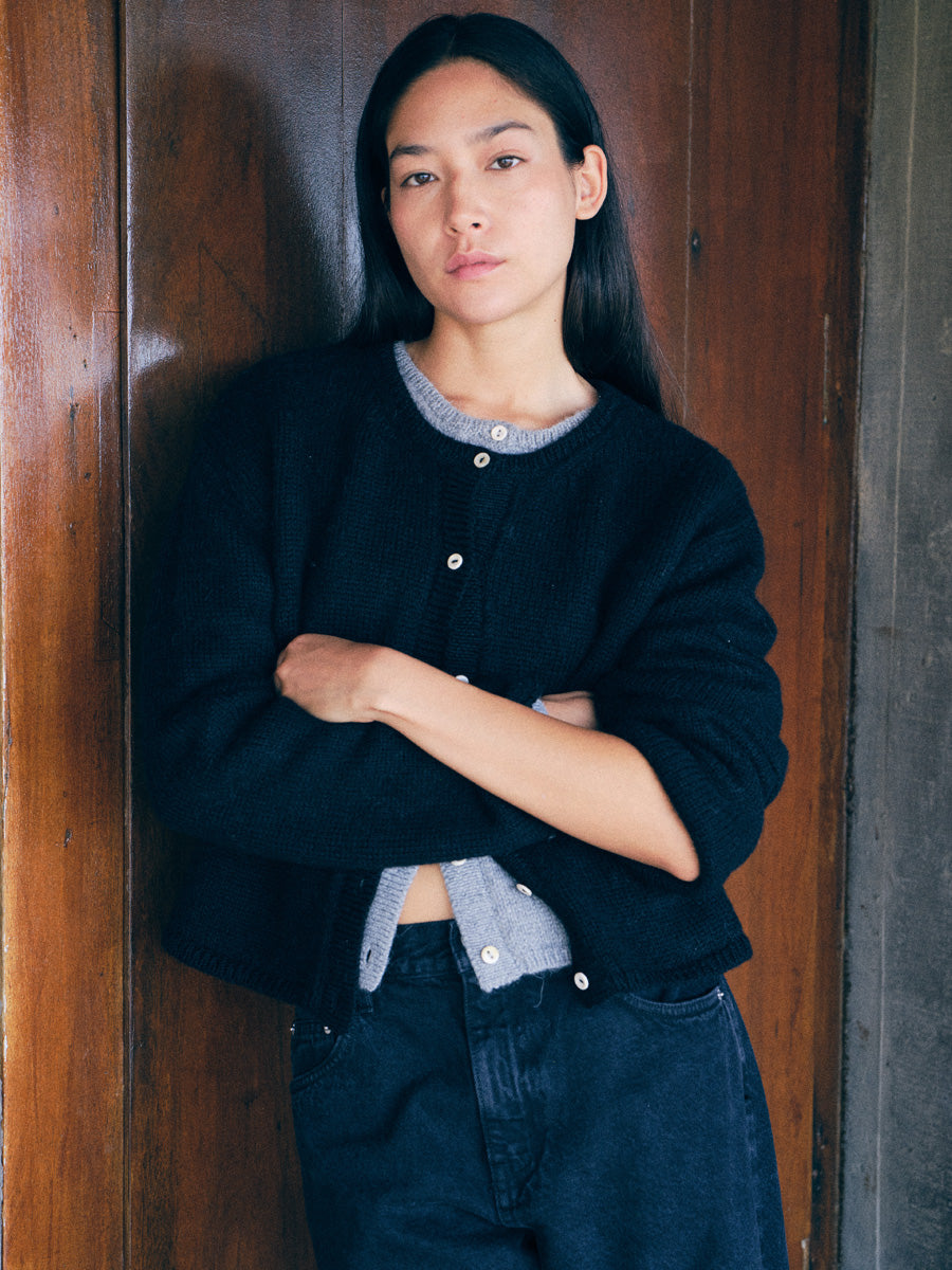 A woman with long dark hair stands with arms crossed, wearing a black knit button-front cardigan over a light gray shirt and dark blue jeans, posed against a rustic wooden door.