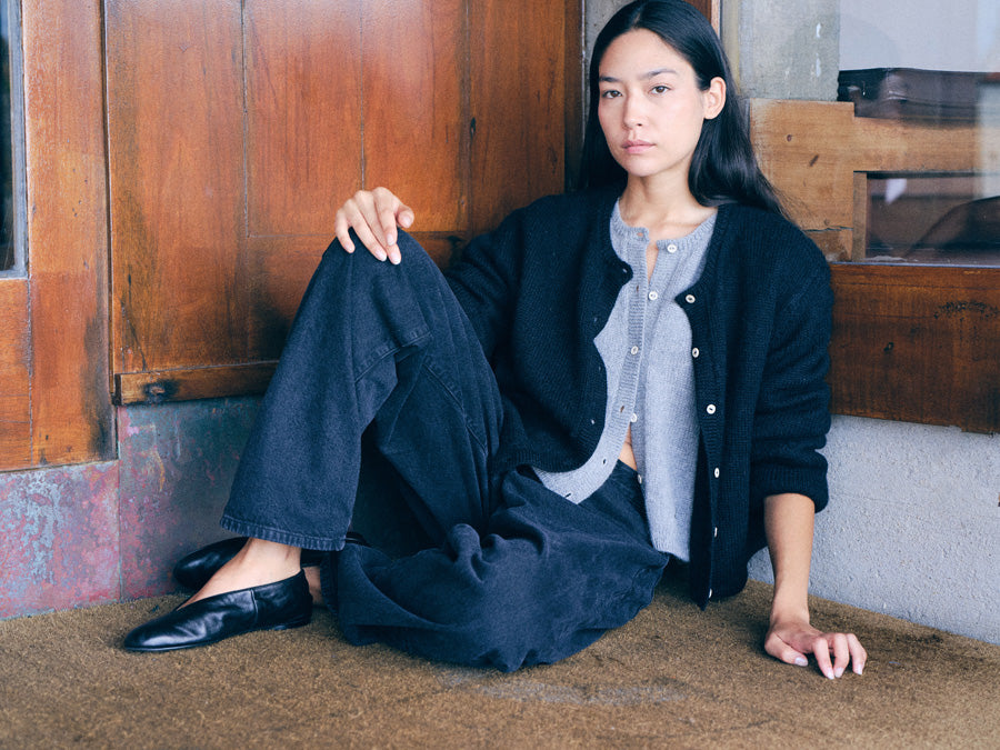 A woman sits on a textured brown floor in front of a rustic wooden door, wearing a black cardigan over a light gray button-up shirt, dark wide-leg jeans, and black leather loafers.