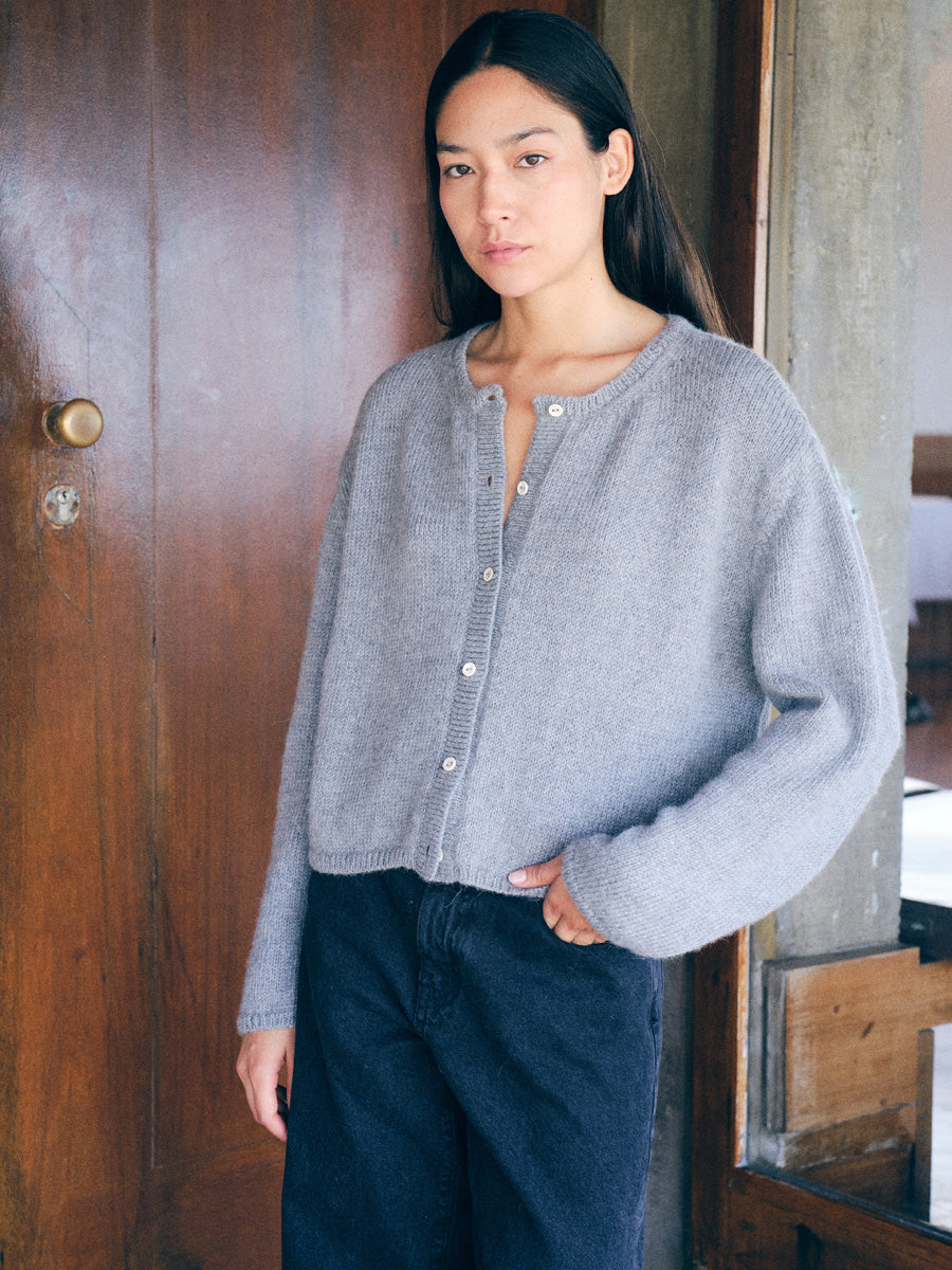 A woman wearing a light gray knit button-front cardigan and dark jeans, standing indoors next to a wooden door.