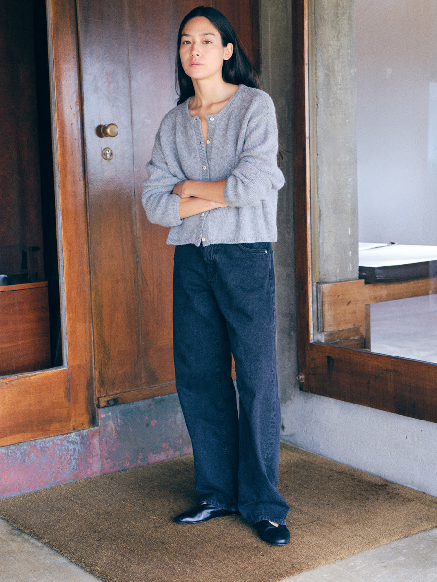 A woman stands on a brown coir doormat in front of a rustic wooden door, wearing a light gray knit cardigan, dark wide-leg jeans, and black slip-on shoes.