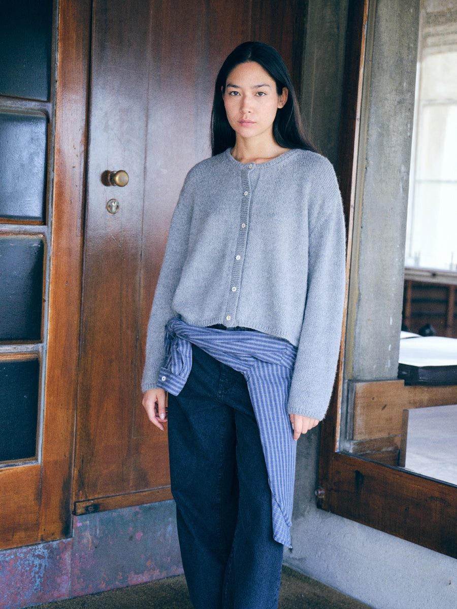 A woman stands indoors wearing a light gray cropped button-front cardigan over a blue striped shirt tied around her waist, paired with dark wide-leg jeans. She is positioned in front of a large wooden door and concrete wall.