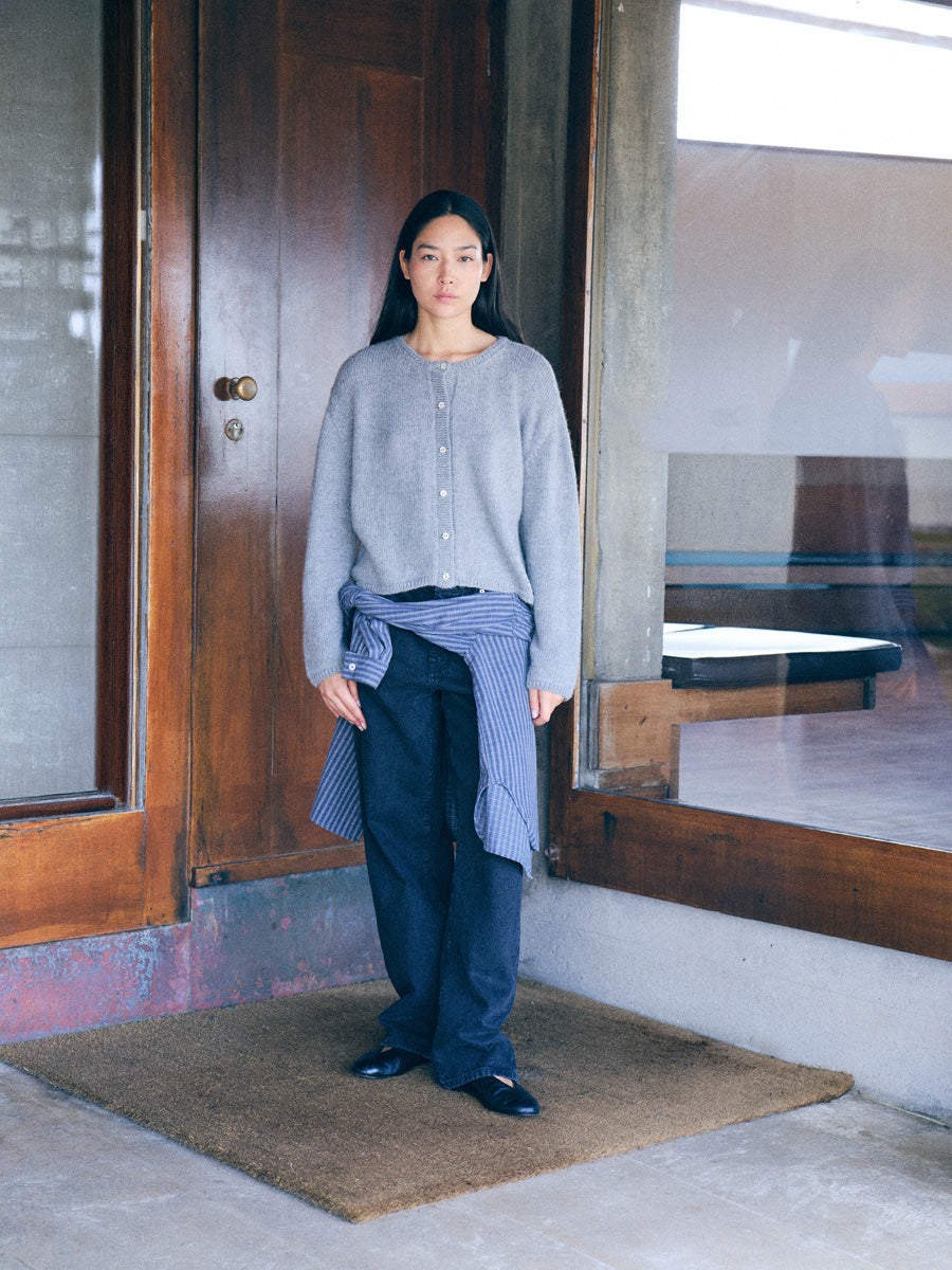 A woman stands on a brown doormat at a wooden building entrance, wearing a light gray knit cardigan, dark wide-leg pants, and a blue striped shirt tied around her waist.